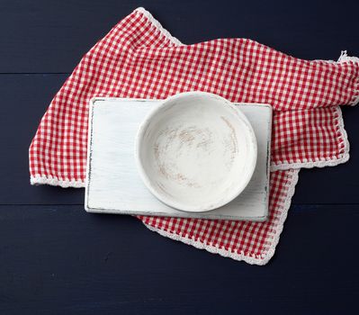 rectangular white cutting kitchen board and empty round plate on a blue wooden background, next to a white red textile towel, top view. Copy space