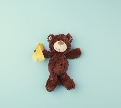 brown teddy bear holds in his paw a yellow ribbon folded in a loop on a blue background. concept of the fight against childhood cancer. problem of suicides and their prevention