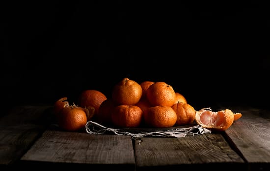 pile of unpeeled round ripe orange mandarin in a plate on a black background, low key