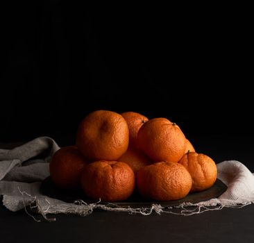 pile of unpeeled round ripe orange mandarin in a plate on a black background, low key