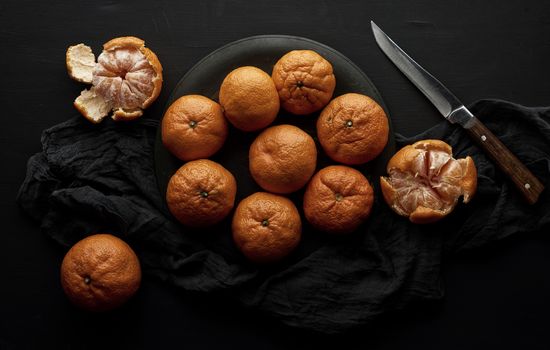 round ripe tangerines in a plate on a black textile napkin and knife, top view, low key