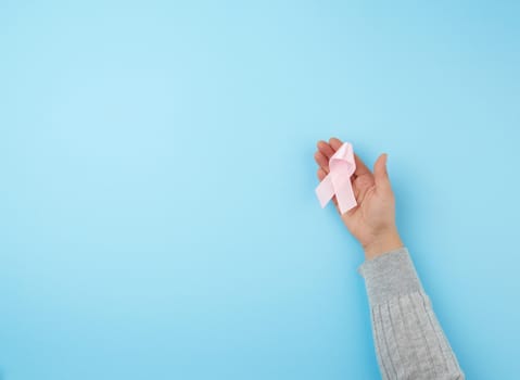 female hand holds a pink bow-shaped ribbon on a blue background. Breast cancer awareness campaign concept.