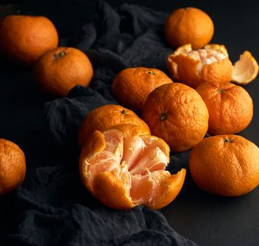 peeled ripe orange mandarin and a bunch of unpeeled round whole fruits on a black napkin, low key