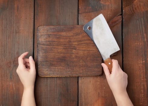 very old empty wooden rectangular cutting board and hands hold a large sharp meat knife, top view, brown wooden background 