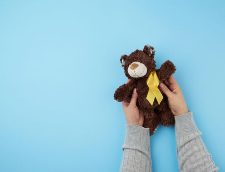 female hands hold a small brown teddy bear which holds in its paw a yellow ribbon folded in a loop on a blue background. concept of the fight against childhood cancer. problem of suicides and their prevention