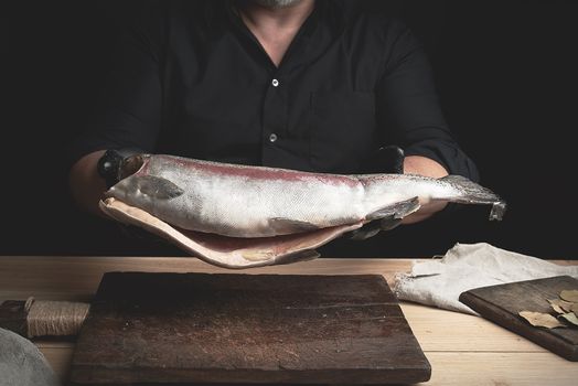 chef in a black shirt and black latex gloves holds a raw carcass of headless salmon fish over a brown wooden cutting board, process of cutting fish