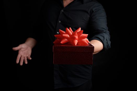 adult man in a black shirt holds a red square box with a knotted bow on a dark background, concept of congratulations, surprise