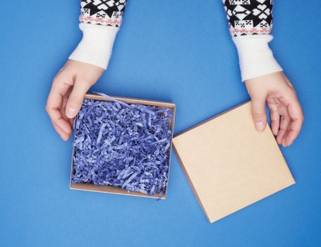 two female hands in a white sweater hold open square brown box on a blue classic background, top view