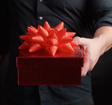 adult man in a black shirt holds a red square box with a knotted bow on a dark background, concept of congratulations, surprise