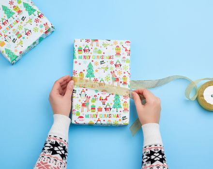 two female hands are wrapping a gift in holiday paper on a blue background, top view, do-it-yourself crafts, festive backdrop