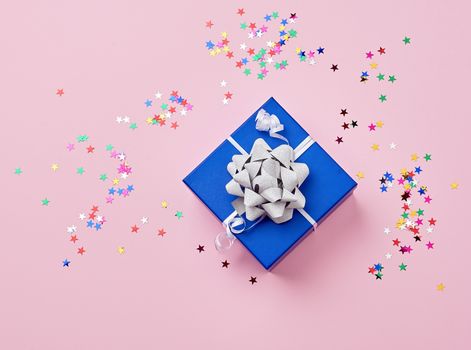 blue cardboard square box tied up with a white ribbon with a shiny bow and scattered multicolored shiny star-shaped confetti on a pink background, holiday backdrop, top view