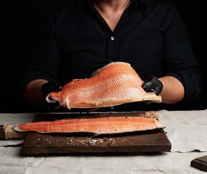 chef in a black shirt and black latex gloves holds a large piece of salmon fish fillet over the table, cooking process, clouse up
