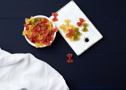 raw pasta in the form of bows in a wooden round plate and white textile napkin on a blue wooden background, top view. Mediterranean food set.