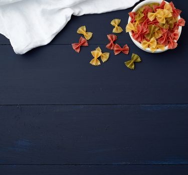 raw pasta in the form of bows in a wooden round plate and white textile napkin on a blue wooden background, top view. Vegetarian nutrition. Italian cuisine