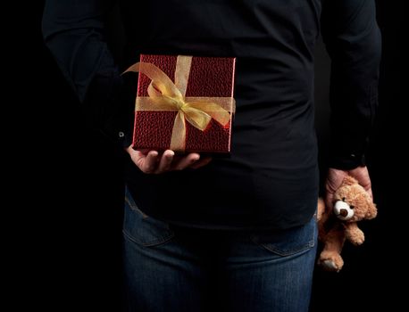 adult man in a black shirt and blue jeans holds a red gift box with a bow behind him, concept of surprise