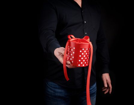 adult man in a black shirt holds in his hand a round red box of polka dots with a red bow on a black background, concept of a holiday greetings, surprise and gratitude
