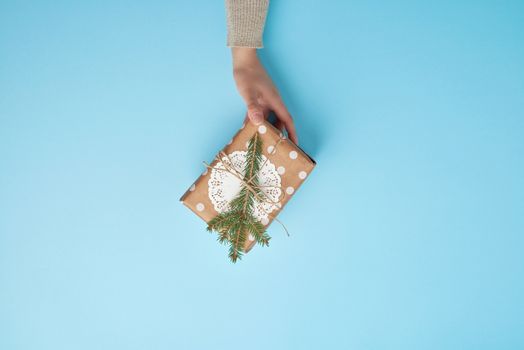 female hand holding a gift wrapped in a brown paper and decorated with a rope and a sprig of spruce on a blue background, top view
