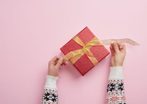 two female hands are holding a red box wrapped in gold ribbon on a pink background, top view. Holiday greetings concept
