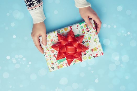 two female hands hold a paper closed box with red bow on a blue background. Festive concept
