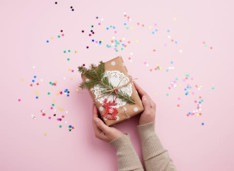 two female hands holding a gift wrapped in paper and decorated with a rope and a sprig of spruce on a pink background, top view