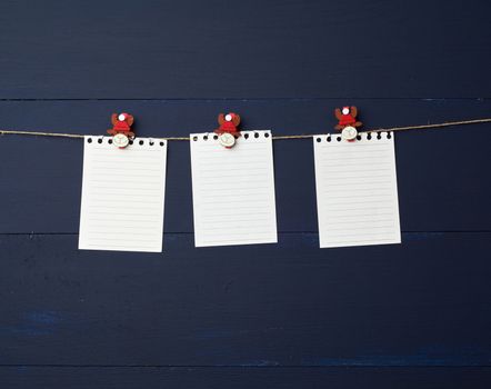 empty white notebook paper sheets hanging on decorative holiday clothespins, blue wooden background. Clean sheet, template. Design element.