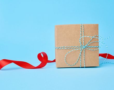 square brown cardboard box for a gift and twisted silk red ribbon on a blue background. Beautiful box gift, great design for any purposes. 