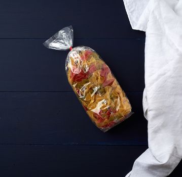 transparent cellophane bag with raw colorful pasta in the form of bows on a blue wooden surface, copy space