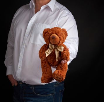 adult man in a white shirt holds a brown teddy bear on a dark background, concept of congratulations on the holiday of Valentine's Day on February 