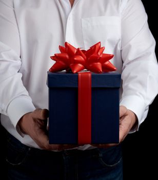 adult man in a white shirt holds a blue gift cardboard box with a red bow, concept of a congratulation on the holiday, anniversary, surprise