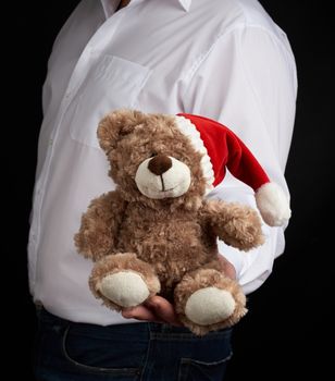 man in a white shirt holds a teddy brown bear in a red cap, Christmas toy