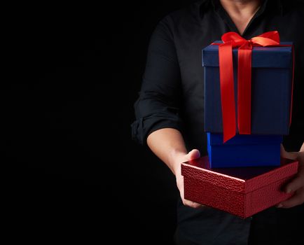 man in a black shirt holds in his hands a stack of paper-wrapped gifts on a black background, concept of holiday greetings, surprise and gift, copy space