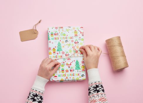 two female hands are wrapping a gift in holiday paper on a pink background, top view, do-it-yourself crafts, festive backdrop