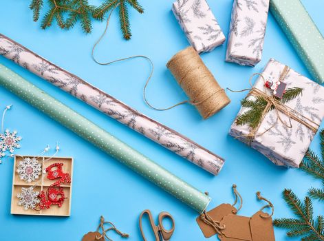 rolls with wrapping paper, brown rope, scissors, decor and a wrapped square box with a gift. DIY gift making process, blue background
