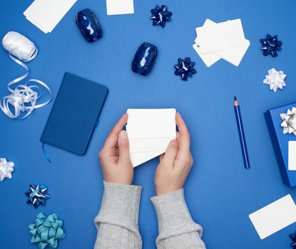 two female hands are holding white paper blank business cards, on a blue background are wrapped gifts and decor, top view