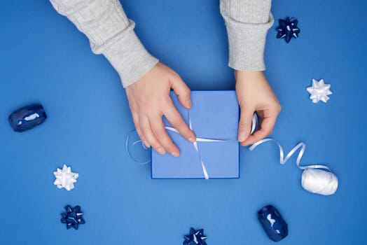 two female hands in a gray sweater hold closed square blue box with a bow on a blue classic background, top view, festive backdrop
