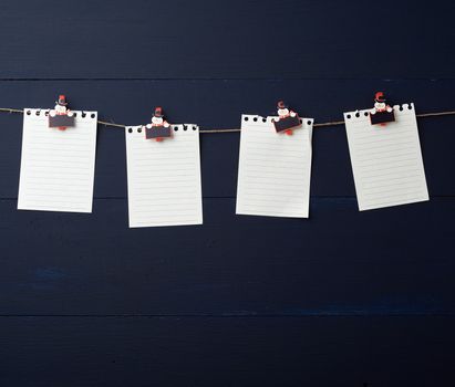 empty white notebook paper sheets hanging on decorative holiday clothespins, blue wooden background. Clean sheet, template. Design element.
