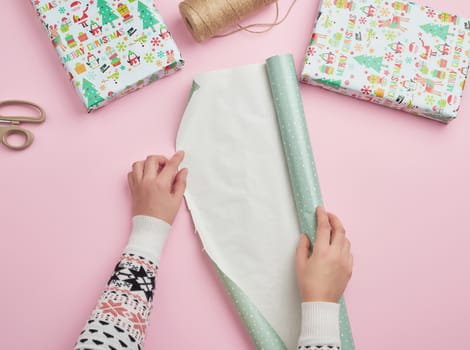 two female hands unfold a roll of wrapping paper, next to two gifts, pink background, preparing surprises