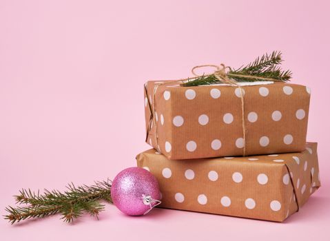 stack of kraft paper wrapped gifts, spruce branch and pink christmas ball on pink background. Christmas greeting concept.