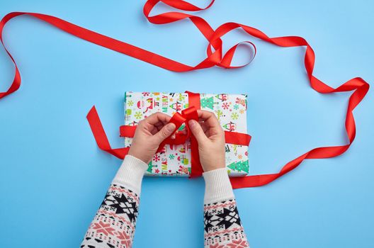two female hands in a sweater knotted a red ribbon on a wrapped Christmas present on a blue background, top view. Festive concept