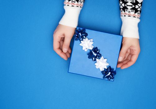 two female hands in a gray sweater hold closed square blue box with a bow on a blue classic background, top view, festive backdrop