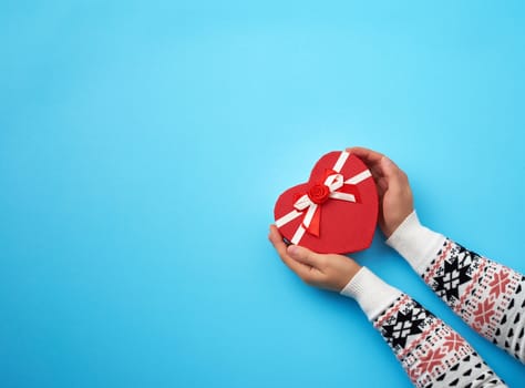 two female hands hold a paper closed red box in the shape of a heart on a dark blue background, festive backdrop, copy space
