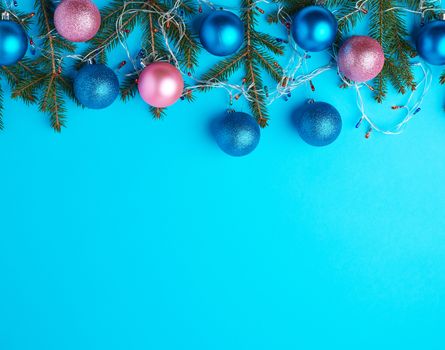 burning multi-colored light bulbs on a garland with a white cable and green twigs, blue and pink balls, blue background, copy space