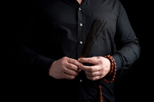 adult man in black clothes holds a stack of incense sticks, dark background