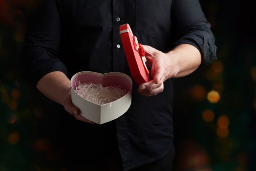 adult man in a black shirt holds a red cardboard box in the form of a heart with a bow on a dark background, concept of holiday poisoning, surprise and a gift