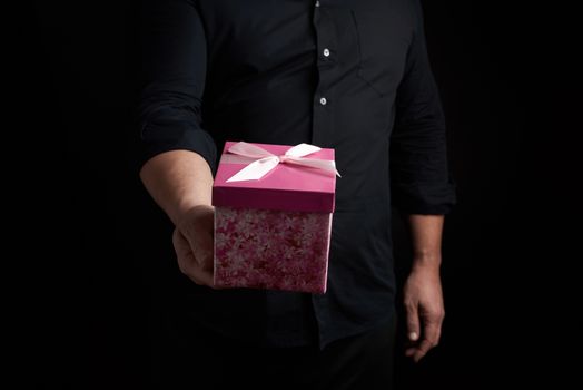 adult man in a black shirt holds a pink square box with a bow tied on a dark background, concept of congratulations, surprise