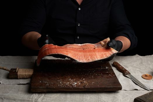 chef in a black shirt and black latex gloves holds a large piece of salmon fish fillet over the table, cooking process, clouse up