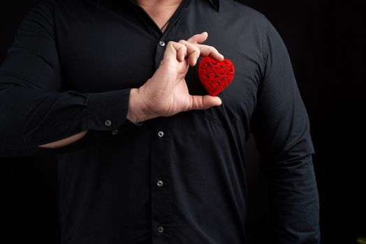 adult man stands on a dark background wearing a black shirt and holding a red carved heart near his chest in his right hand, concept of love