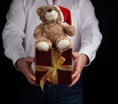 adult man in a white shirt holds a red square box tied with a golden ribbon and brown teddy bear, Merry Christmas and Happy New Year greetings concept