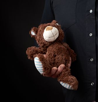 adult man in a black shirt holds a brown teddy bear on a dark background, concept of congratulations on the holiday of Valentine's Day on February 14, low key