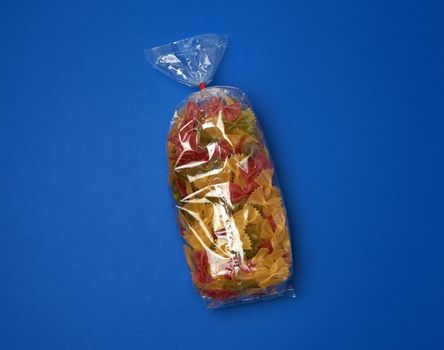 transparent cellophane bag with raw colorful pasta in the form of bows on a blue surface, copy space